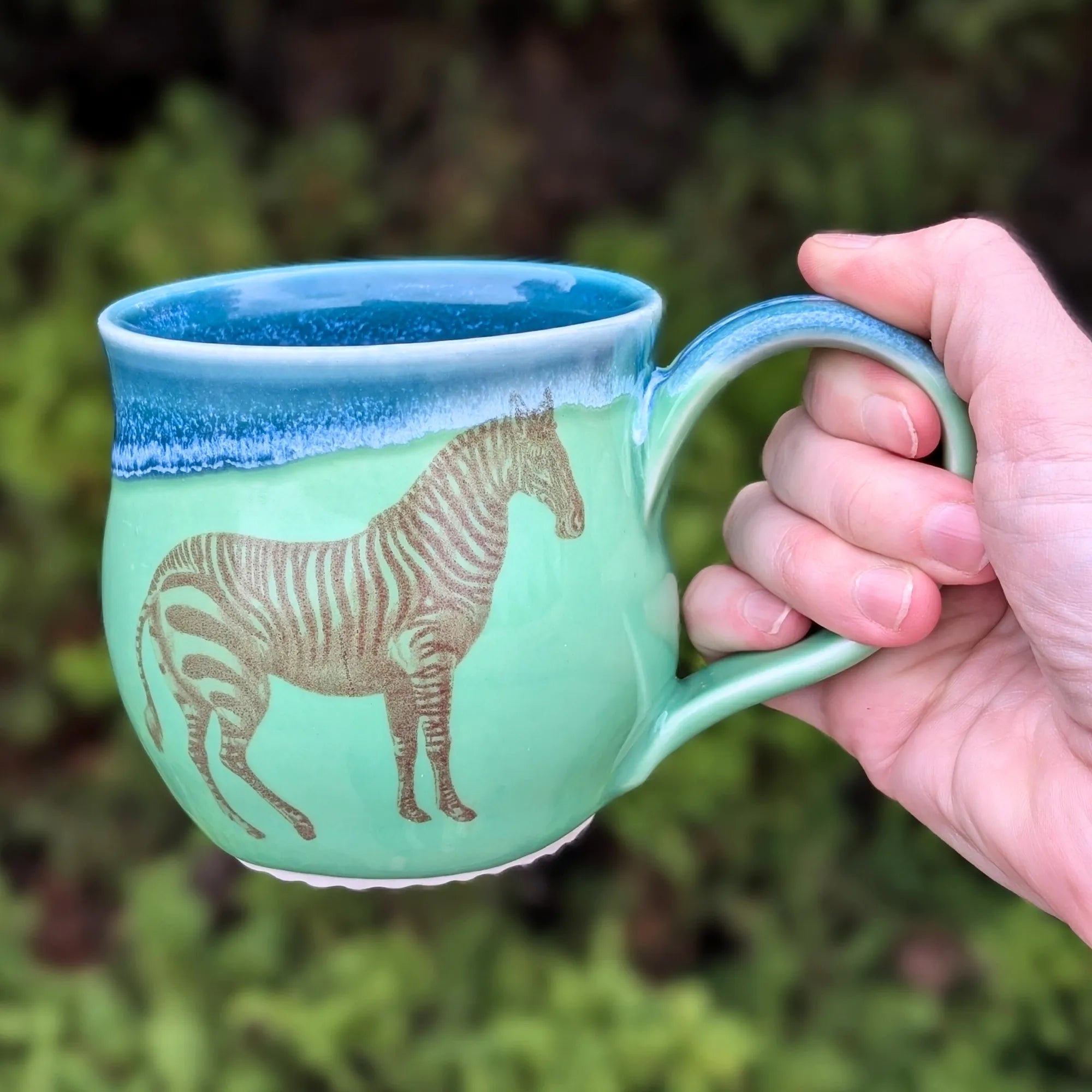 Hand holding a green mug with a zebra design against a blurred natural background